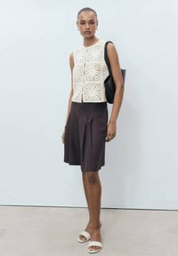 Crochet top in cream with square patterns, paired with dark brown pleated skirt and white slide sandals. Accessory: black shoulder bag.