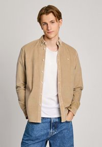 Lichtbeige corduroy overhemd met een button-down kraag, gedragen over een wit T-shirt; gecombineerd met blauwe jeans. Eenvoudig borduurdetail op het overhemd.