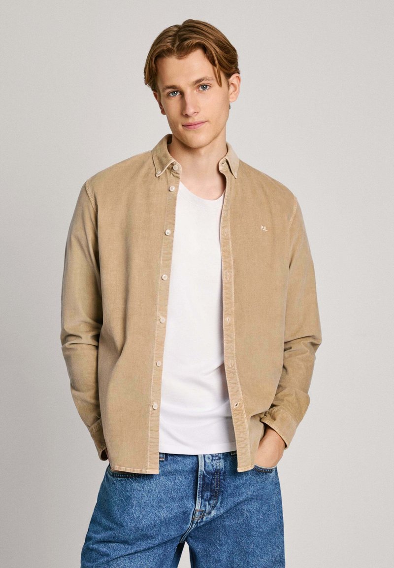 Lichtbeige corduroy overhemd met een button-down kraag, gedragen over een wit T-shirt; gecombineerd met blauwe jeans. Eenvoudig borduurdetail op het overhemd.