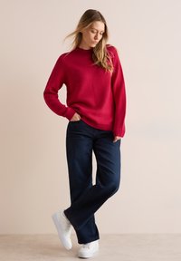 Rode gestructureerde breipullover met een hoge hals, gecombineerd met donkerblauwe wijde jeans en witte platform sneakers. Eenvoudige en casual outfit.
