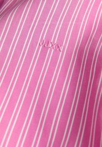 Lyserød stribet stofskjorte med tynde hvide striber og et broderet logo i matchende lyserød. Glat tekstur, afslappet design.