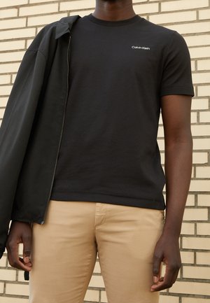 Man die een zwart Calvin Klein T-shirt draagt, beige broek, en een zwart jasje over één schouder houdt tegen een achtergrond van een bakstenen muur.