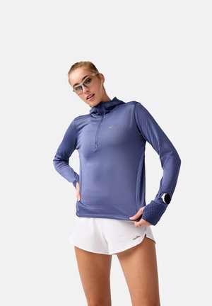 Haut de sport violet à manches longues avec col zippé, tissu texturé et trous pour les pouces. Associé à un short blanc. Lunettes de soleil et montre de fitness incluses.