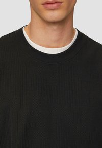 Zwarte sweater met textuur en een geribbelde halslijn, gelaagd over een wit T-shirt met ronde hals, met een close-up van de stofdetails.