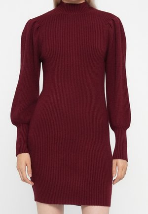 Femme portant une robe tricotée bordeaux ajustée à manches longues côtelées avec des épaules bouffantes et un col montant, debout devant un fond uni.