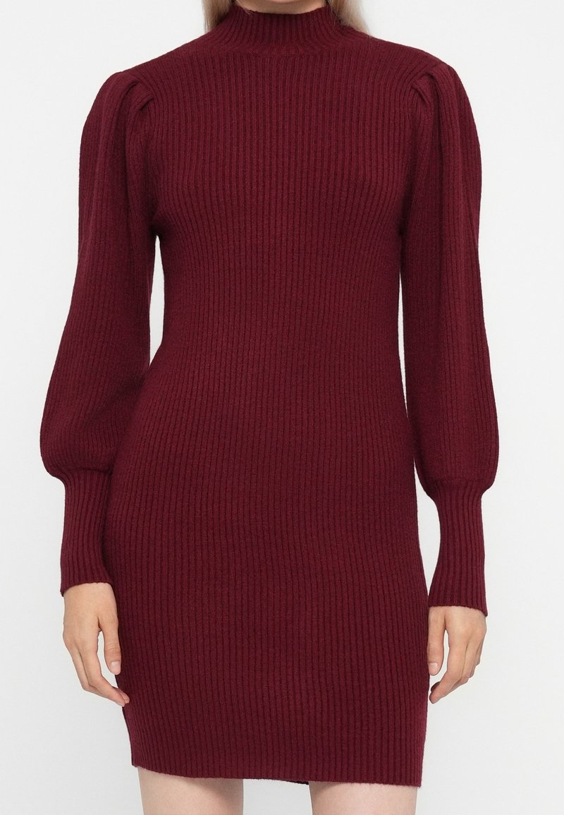 Femme portant une robe tricotée bordeaux ajustée à manches longues côtelées avec des épaules bouffantes et un col montant, debout devant un fond uni.