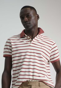 Camisa polo de algodón con rayas rojas horizontales sobre un fondo crema, con un cuello rojo en contraste y mangas cortas.