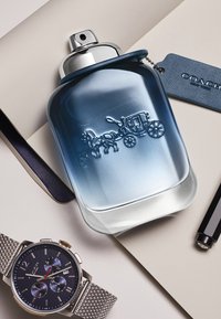 Bottiglia di profumo in vetro con un design sfumato che va dal blu al trasparente, caratterizzata da un cavallo e una carrozza in rilievo. Accompagnata da un orologio e una penna.