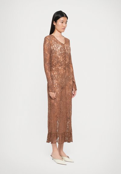 Robe en dentelle marron avec des manches longues, décolleté en V, détails froncés à la taille et ourlet évasé. Le modèle porte des mules blanches à bout pointu.