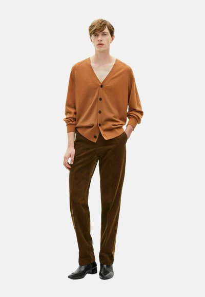 Brauner Cardigan mit V-Ausschnitt, lockerer Passform und schwarzen Knöpfen, kombiniert mit dunkelgrünen Cordhosen und schwarzem Schuhwerk.