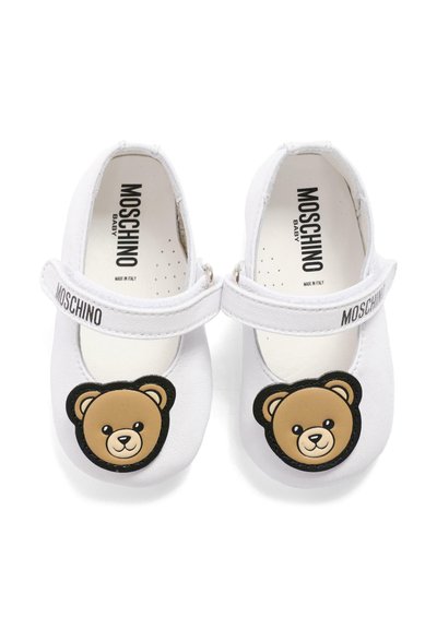 Chaussures de bébé blanches avec des visages d'ours en peluche sur les bouts des orteils et le nom de la marque "Moschino" sur les brides et les semelles intérieures.