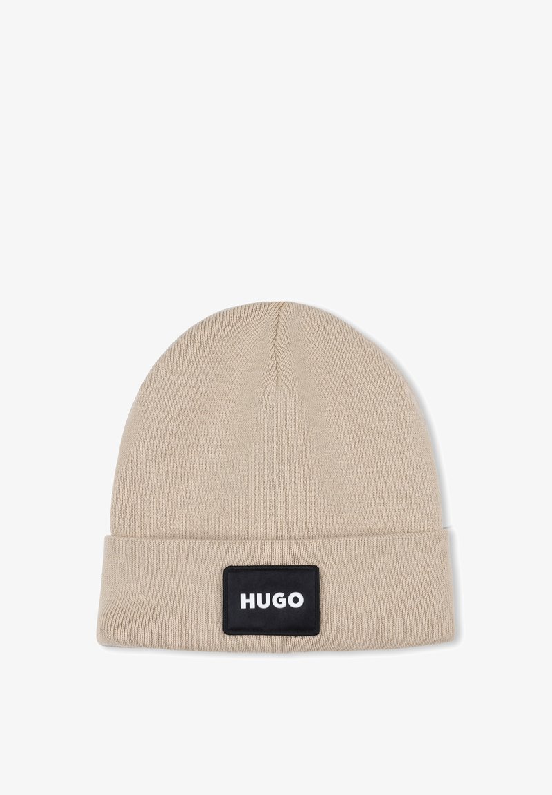 Bonnet en tricot beige avec un revers. Il présente une patte rectangulaire noire avec le texte blanc "HUGO". Texture lisse, design simple.