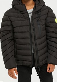 Veste doudoune noire avec capuche, présentant un design matelassé, une poche poitrine zippée et un patch logo en accent sur la manche gauche.