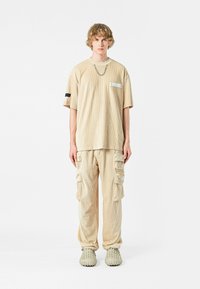Camiseta oversized de corduroy beige claro con un bolsillo en el pecho, combinada con pantalones cargo a juego que cuentan con múltiples bolsillos. Calzado grueso gris.