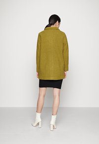 Olivgrön texturerad kappa med krage, något oversized passform, och svart ribbad kjol, kombinerad med vita klackade ankelskors.
