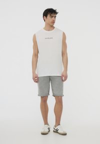 Canottiera bianca senza maniche con testo stampato nero, abbinata a pantaloni corti di jeans grigi e sneakers bianche con strisce nere. Look semplice e casual.