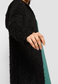 Cardigan negro de felpa con capucha, que presenta una textura suave y un corte abierto, combinado con una prenda verde debajo. Mano extendida.