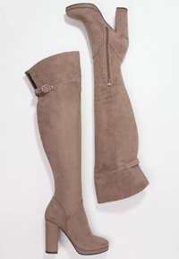 Botas de ante beige hasta la rodilla con tacones gruesos, cremallera lateral y detalles de hebilla en el muslo. Textura suave y forma ajustada.