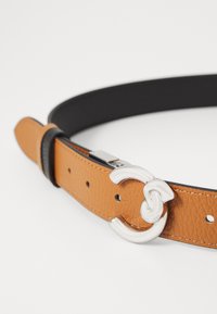 Cinturón de cuero en color marrón claro con un acabado texturizado, que presenta una hebilla circular de metal plateado y un interior negro. Diseño delgado con agujeros ajustables.