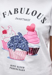 Λευκό πουκάμισο με τρία διακοσμημένα cupcakes: ένα με μπλε παγιέτες, ένα με ροζ καρδιά και ένα με κεράσι, με κείμενο για τη γλυκύτητα.