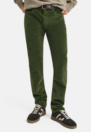 Homme portant un pantalon en velours côtelé vert, un pull beige et des baskets noires avec des lacets et des rayures blanches, se tenant les mains dans les poches.