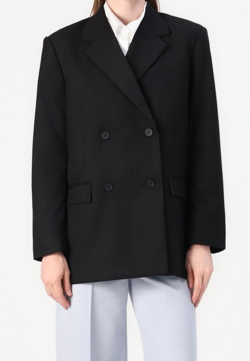 Blazer noir croisé en laine, avec un col pointu, cinq boutons et deux poches latérales. Associé à un pantalon bleu clair.