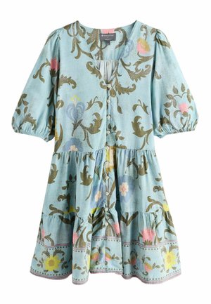 Woman in a light blue floral dress holding a gray textured coat, standing outdoors with trees and grass in the background.