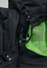 Svart ryggsäck med strukturerad tyg, en grön meshnäta ficka och svarta dragkedjor. Inkluderar plastdetaljer och justerbara remmar.