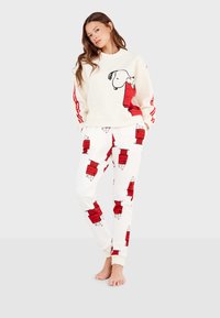 Ensemble sweat-shirt et pantalon crème avec un design de chien cartoon rouge ; les pantalons présentent des impressions répétées de chemises rouges. Tissu doux, coupe décontractée.