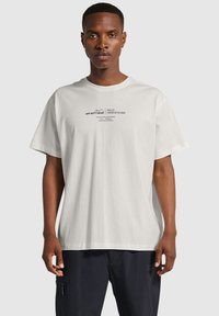 Vit kortärmad t-shirt i bomull, med en grafisk tryck i svart text på framsidan. Klassisk rundad halsringning och lös passform.