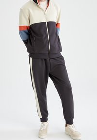 Veste de course pour homme et pantalon en noir, crème, bleu et orange. La veste dispose de poches zippées et de poignets côtelés. Tissu lisse et extensible.