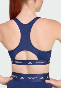 Modra športna športna modrček z racerback obliko, teksturirano tkanino in kontrastnim logotipnim trakcem z oznako "TECHFIT" ob dnu.