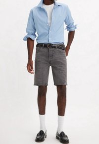 Hombre con camisa de botones azul claro con las mangas arremangadas, camiseta blanca, pantalones cortos de mezclilla grises, cinturón trenzado negro, calcetines blancos y mocasines negros.