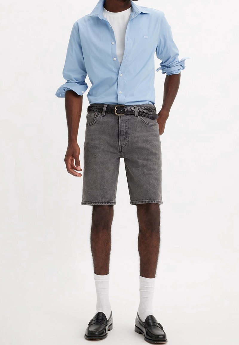 Hombre con camisa de botones azul claro con las mangas arremangadas, camiseta blanca, pantalones cortos de mezclilla grises, cinturón trenzado negro, calcetines blancos y mocasines negros.
