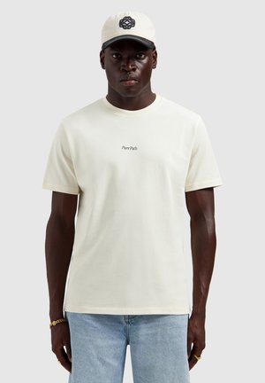 Man draagt een witte "Pure Path" T-shirt, lichtblauwe spijkerbroek, witte pet met zwart logo, gouden armband en ringen, staand tegen een effen achtergrond.
