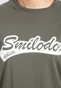 Camiseta de color verde oliva hecha de algodón. Presenta un gran texto gráfico en blanco que dice "Smilodon" con la palabra "Atleta" abajo en un diseño curvado.