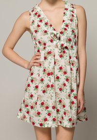 Kukallinen mekko, jossa on V-mallinen kaula-aukko, hihaton muotoilu ja ryppyvyötärö, jossa on solmusoikeus. Väreinä valkoinen, punaiset ruusut ja vihreät lehdet.