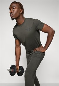 Olijfgroen sportshirt en -broek, met een subtiele logo. Het model houdt een zwarte dumbbell vast en toont een workout-houding.