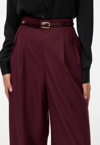 Pantaloni ampi color bordeaux con una texture liscia, caratterizzati da pieghe e una cintura lucida color marrone scuro con una fibbia dorata in rilievo. Abbinato a una blusa nera.