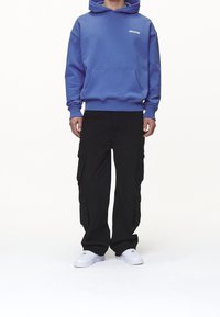 Hombre con sudadera azul que tiene el texto "PEGADOR", pantalones cargo negros y zapatillas blancas, de pie contra un fondo blanco.