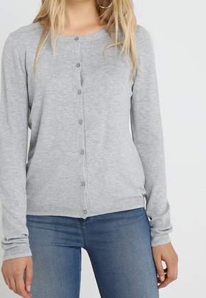 Femme portant un cardigan gris clair boutonné à manches longues et un jean bleu, debout devant un fond clair uni.