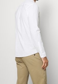 Camicia bianca a maniche lunghe in misto lino, con polsini con bottone, piega sul retro e vestibilità aderente, indossata con pantaloni khaki.