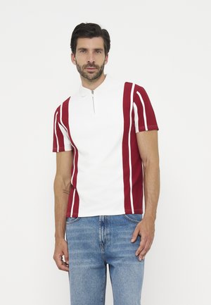 Uomo che indossa una polo bianca con righe verticali rosse e jeans azzurri chiari, in piedi davanti a uno sfondo bianco semplice.