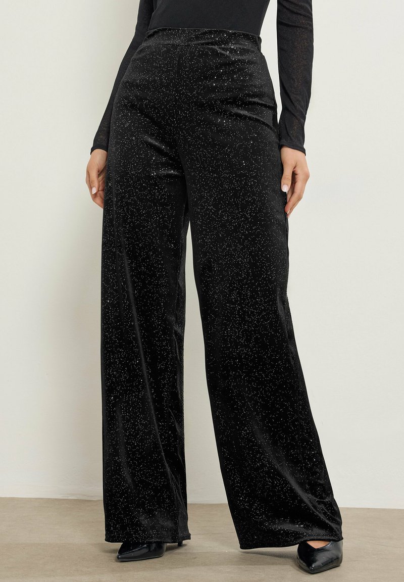 Zwarte fluwelen broek met wijde pijpen en een zachte textuur, voorzien van verspreide zilveren spikkels voor een sprankeling. Glad tailleband, volledige lengte tot de vloer.