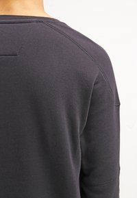 Sudadera negra con una textura suave, cuello redondo y mangas raglán. Presenta una costura en el hombro discreta y un pequeño bolsillo de parche.
