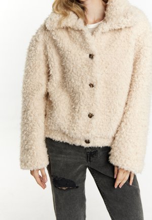 Vrouw draagt een crèmekleurige, met knopen sluitende, getextureerde fleecejas, gecombineerd met zwarte jeans met scheuren, tegen een effen achtergrond.