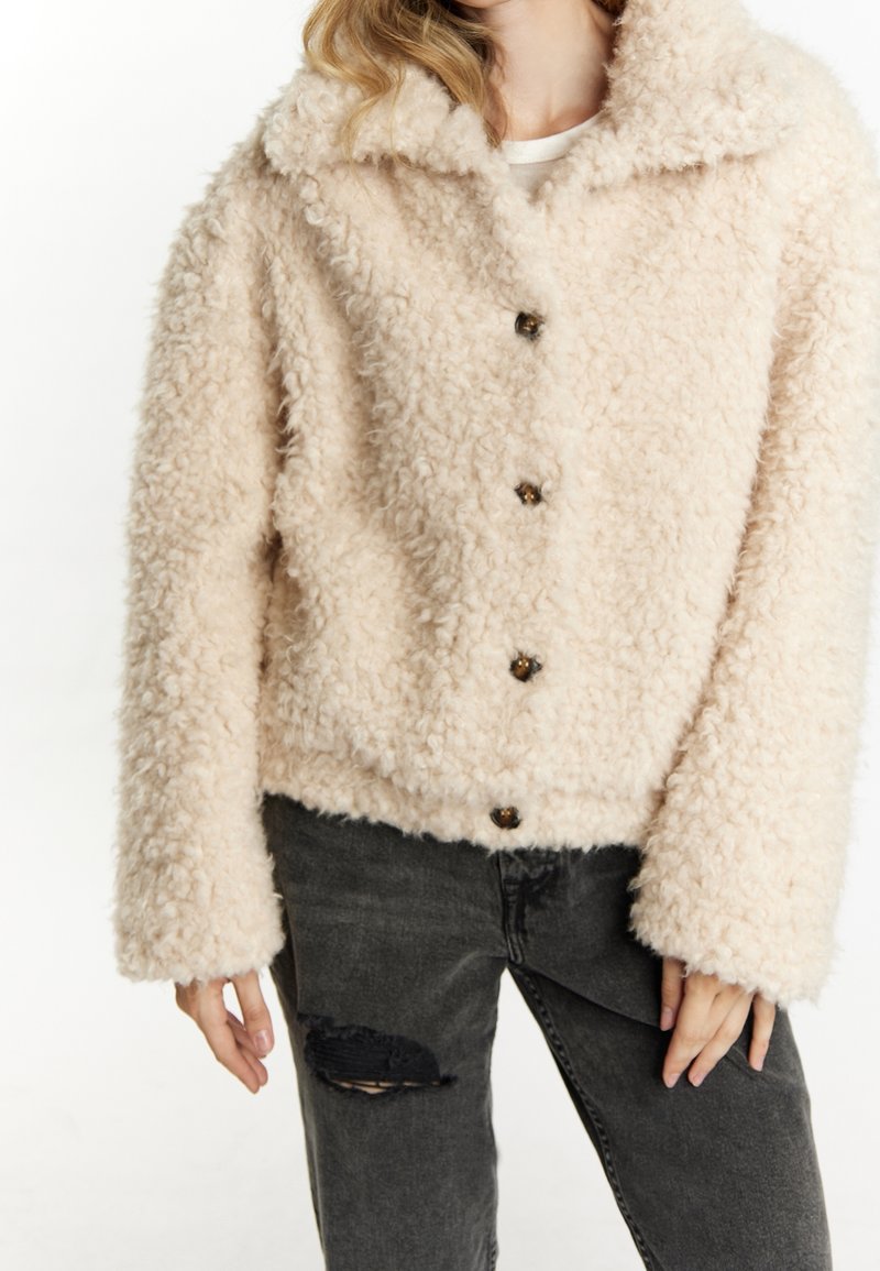 Femme portant une veste polaire texturée crème avec boutons, associée à un jean noir déchiré, sur un fond uni.
