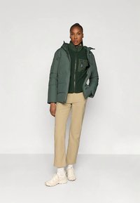 Egy nő áll egy sötétzöld pufertakaró kabátban, zöld cipzáras felsőben, bézs nadrágban, fehér zoknikban és fehér sportcipőben, egy sima világos háttér előtt.