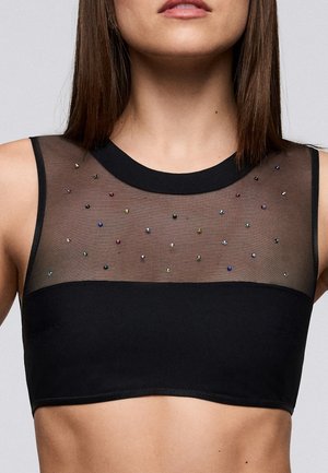 Vrouw draagt een zwarte mouwloze crop top met een doorzichtig bovendeel versierd met kleine, kleurrijke kraaltjes.