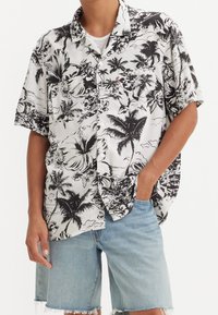 Camicia a maniche corte bianca e nera con colletto, decorata con motivi di ibisco e palme, chiusura con bottoni e vestibilità comoda.